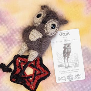 May include: A fuzzy dark gray crocheted owl doll with large black eyes and beige accents, positioned over a black and red crocheted pentagram. Beside it lies a white rectangular card labeled STOLAS with an owl illustration and text.