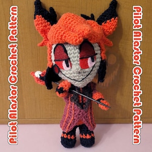Puede incluir: Una muñeca de ganchillo hecha a mano de un personaje con cabello naranja brillante, orejas negras y cara pálida. La muñeca viste un abrigo rojo oscuro y negro con detalles en naranja y sostiene un bastón negro. El texto lateral dice Pilot Alastor Crochet Pattern.