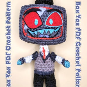 May include: A crocheted doll featuring a television-shaped head with red eyes, a blue mouth, and a black top hat. The body wears a grey vest, white shirt, red tie, and black trousers. The text "Box Vox PDF Crochet Pattern" appears on both sides.