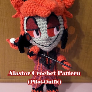Puede incluir: Muñeca de ganchillo hecha a mano de un personaje con pelo rojo, detalles negros y un atuendo rojo y negro. La muñeca tiene una cara sonriente y sostiene un bastón negro. El texto en la imagen dice "Alastor Crochet Pattern (Pilot-Outfit)".