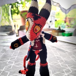 May include: A handcrafted crochet doll featuring a red and white head with tall, black and white striped horns and yellow eyes. The figure wears a dark blue coat over a red torso with a white cross symbol, black pants, and a red tail with a pointed tip.