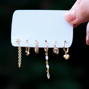 Puede incluir: Un conjunto de seis pendientes dorados exhibidos en una tarjeta blanca. Los pendientes incluyen varios diseños, como una cadena, aros y un dije en forma de corazón. Los pendientes son sostenidos por una mano contra un fondo verde borroso.