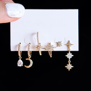 Peut inclure: Un ensemble de boucles d'oreilles dorées avec des motifs célestes, dont des étoiles et des croissants de lune, présentées sur une carte blanche sur fond noir. Les boucles d'oreilles comportent de petits ornements scintillants.