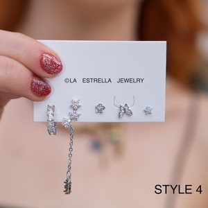 May include: A white card with the text "OLA ESTRELLA JEWELRY" and "STYLE 4" on it. The card displays a set of six silver earrings with clear cubic zirconia stones. The earrings include a hoop, a dangle with a chain, a butterfly, two flowers, and a single stone.