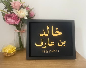 May include: A black framed sign with gold Arabic calligraphy that reads "Khalid bin Aaraf 1444 H". The sign is on a wooden surface with a vase of pink flowers and a glass bowl with a yellow flower.