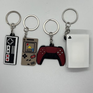 Peut inclure: Quatre porte-clés représentant différents contrôleurs de jeux vidéo. Les porte-clés sont argentés et présentent une manette PlayStation rouge et noire, une Game Boy marron et jaune, une manette Nintendo blanche et noire et une console PlayStation 5 blanche.