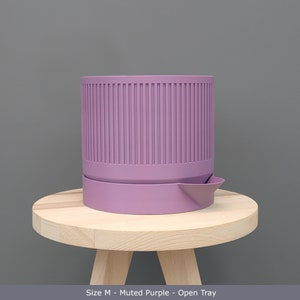 Peut inclure: Un pot de fleurs de couleur mauve avec un design côtelé et un plateau ouvert. Le pot est posé sur un tabouret en bois.