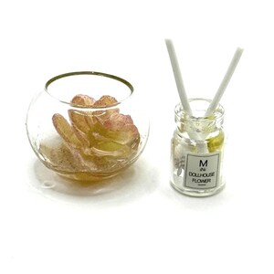 May include: A miniature dollhouse decor set. It includes a clear glass bowl with yellow and pink petals and a small glass bottle with white sticks. The bottle has a label that reads "M IN DOLLHOUSE FLOWER".