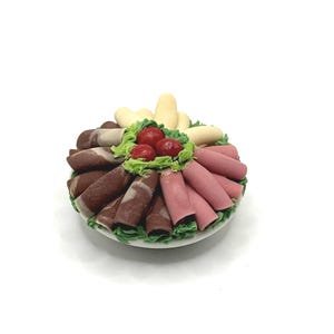 May include: A miniature food platter featuring an assortment of deli meats, such as dark brown, light pink, and white rolls, presented on a bed of green lettuce. Red cherry tomatoes are in the center. The platter is on a white plate.