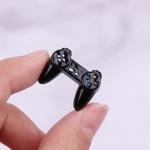 Peut inclure: Une petite manette de jeu noire avec des boutons colorés. La manette a un design classique avec deux joysticks et un pavé directionnel. Les boutons sont bleus, verts, jaunes et rouges. La manette est tenue entre deux doigts.