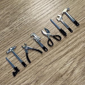 Peut inclure: Une collection de petits outils argentés, dont un marteau, une scie, une pince, une clé et un tournevis, avec des manches noirs. Les outils sont disposés en diagonale sur une surface en bois.