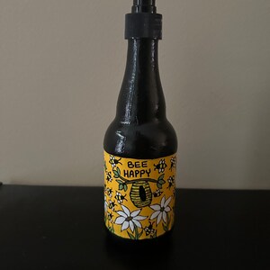 May include: A black glass bottle with a pump dispenser, decorated with a yellow label featuring a beehive, bees, flowers, and the text "Bee Happy".