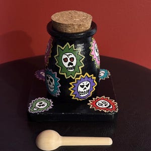 May include: A black jar with a cork stopper, decorated with colorful skull designs. The jar sits on a matching black base, accompanied by a small wooden spoon. The skulls are in various colors, including green, pink, and purple.