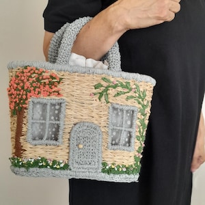 May include: A straw-colored handbag with a house design. The bag features a gray roof and door, with pink and green embroidered details of flowers and foliage. The handles are gray, and the bag is held by a person wearing a black dress.