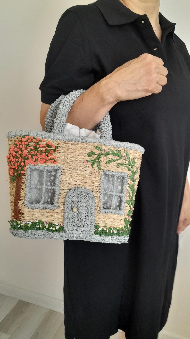 May include: A straw-colored handbag with a house design. The bag features a gray roof and door, with pink and green embroidered details of flowers and foliage. The handles are gray, and the bag is held by a person wearing a black dress.