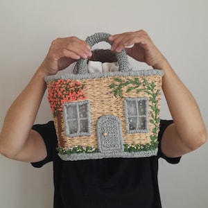 May include: A whimsical house-shaped handbag with a woven straw body, gray crocheted trim, and a matching handle. The facade features embroidered details like windows, a door, and floral accents in pink and green.