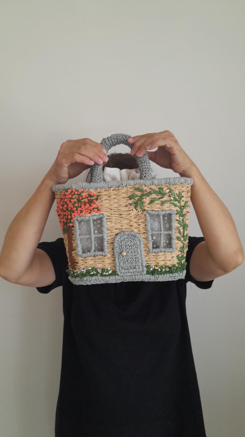 May include: A whimsical house-shaped handbag with a woven straw body, gray crocheted trim, and a matching handle. The facade features embroidered details like windows, a door, and floral accents in pink and green.