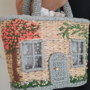 May include: Woven straw handbag with a house design. The bag features a gray handle and trim, with embroidered details of a tree, flowers, and windows. The windows have white dotted curtains. The door has a wooden knob.