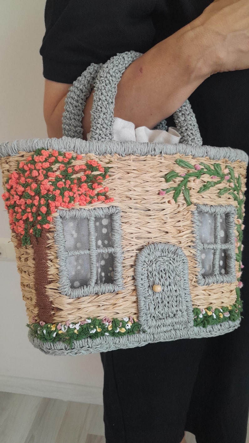 May include: Woven straw handbag with a house design. The bag features a gray handle and trim, with embroidered details of a tree, flowers, and windows. The windows have white dotted curtains. The door has a wooden knob.