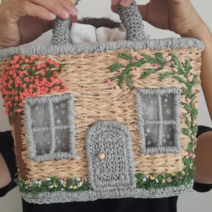 May include: A straw-woven handbag shaped like a house, featuring a gray handle and trim. The house design includes gray windows, a door, and floral accents in pink and green, creating a whimsical cottagecore aesthetic.
