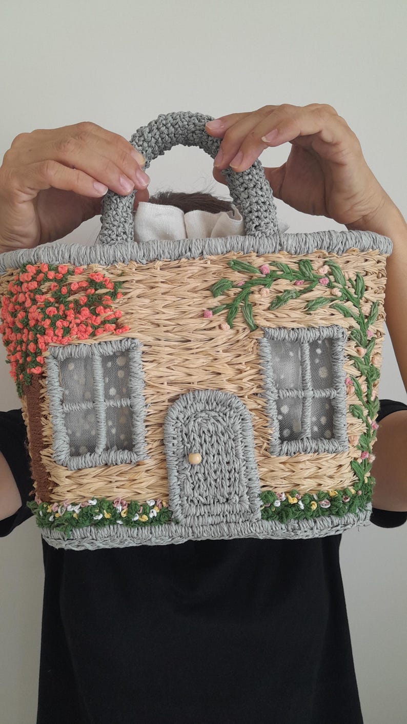 May include: A straw-woven handbag shaped like a house, featuring a gray handle and trim. The house design includes gray windows, a door, and floral accents in pink and green, creating a whimsical cottagecore aesthetic.