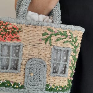 May include: A straw-woven handbag shaped like a cottage, featuring a gray crocheted handle and trim. The bag is adorned with floral and vine embellishments, including pink and green accents, and has a small door and windows.