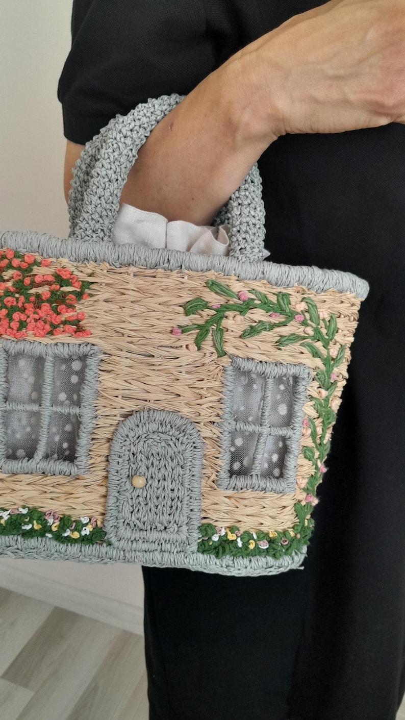 May include: A straw-woven handbag shaped like a cottage, featuring a gray crocheted handle and trim. The bag is adorned with floral and vine embellishments, including pink and green accents, and has a small door and windows.