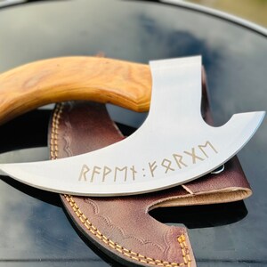 Könnte beinhalten: Eine gebogene Axt aus Edelstahl mit einem Holzstiel. Die Axt hat eine Lederscheide und ist mit dem Text "RFPMT: FARXM" graviert.