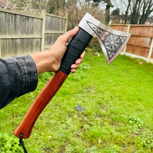 May include: A hand holds a wooden-handled axe with a stainless steel head. The head is engraved with a compass design and a Celtic knot. The axe has a black leather wrap around the handle.
