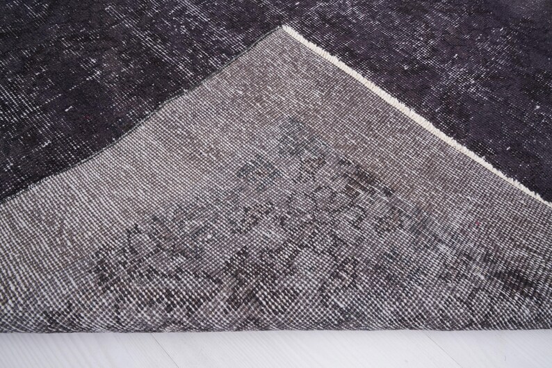 Puede incluir: Una secci&oacute;n de una alfombra gris oscuro y negra con una apariencia desgastada y vintage. La alfombra tiene un patr&oacute;n geom&eacute;trico y un borde blanco. La textura es visible, lo que sugiere un material tejido. La alfombra est&aacute; parcialmente enrollada, revelando la parte inferior.