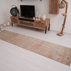 May include: A long, rectangular, light pink and beige runner rug with a faded floral pattern. The rug is laid out on a white wood floor in a living room setting.