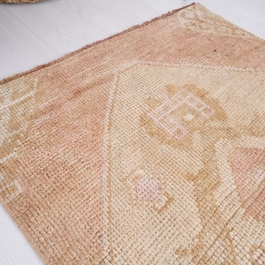 May include: A light brown rug with a faded pink and white geometric pattern. The rug has a distressed look and is made of a soft, woven material.