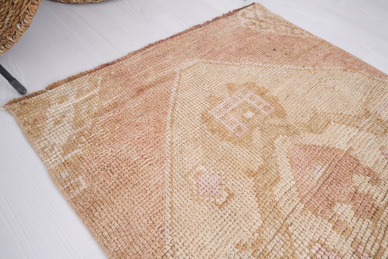 May include: A light brown rug with a faded pink and white geometric pattern. The rug has a distressed look and is made of a soft, woven material.