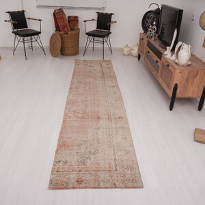 May include: A light pink and cream colored vintage runner rug with a floral pattern. The rug is laid out on a white wood floor in a room with furniture.