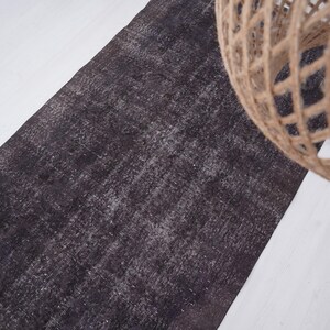 Puede incluir: Una alfombra rectangular gris oscuro con un aspecto desgastado y texturizado. La alfombra est&aacute; colocada sobre una superficie blanca, con un elemento decorativo esf&eacute;rico tejido en la esquina superior derecha.