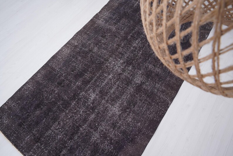 Puede incluir: Una alfombra rectangular gris oscuro con un aspecto desgastado y texturizado. La alfombra est&aacute; colocada sobre una superficie blanca, con un elemento decorativo esf&eacute;rico tejido en la esquina superior derecha.
