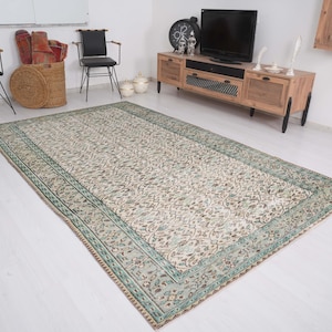 May include: Rectangular area rug with a light beige field and a detailed floral pattern. The rug has a border in shades of green and brown. The rug is placed on a white floor.
