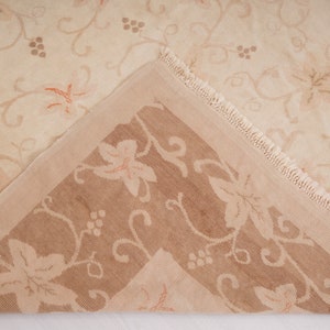 May include: A close-up of a beige and brown patterned rug with a fringe border. The rug has a floral design with a repeating pattern of leaves and grapes.