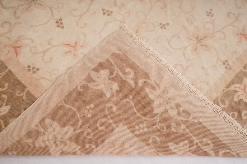 May include: A close-up of a beige and brown patterned rug with a fringe border. The rug has a floral design with a repeating pattern of leaves and grapes.