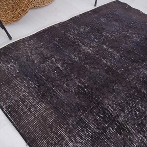 Puede incluir: Una alfombra negra desgastada con reflejos blancos y un borde blanco. La alfombra tiene un aspecto vintage y desgastado. Una cesta tejida y patas de metal negro son visibles en el fondo.