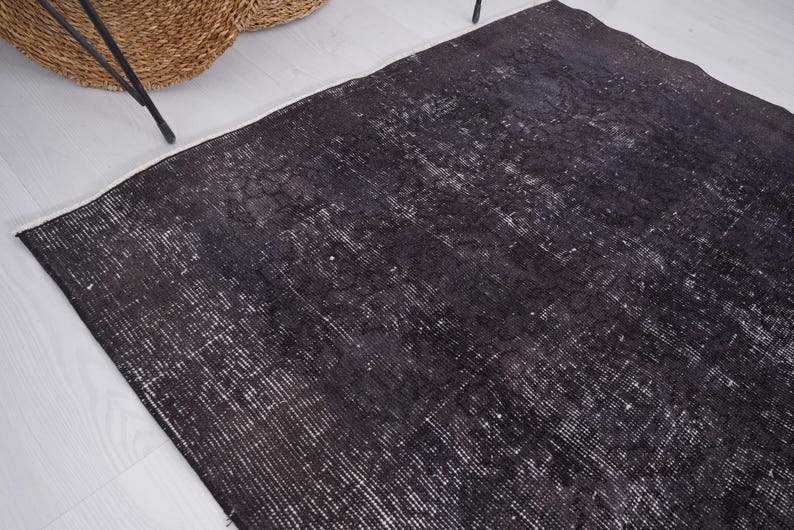Puede incluir: Una alfombra negra desgastada con reflejos blancos y un borde blanco. La alfombra tiene un aspecto vintage y desgastado. Una cesta tejida y patas de metal negro son visibles en el fondo.