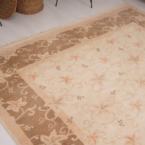 May include: A beige area rug with a brown border. The rug has a floral pattern with a repeating vine design. The border has a similar floral pattern.