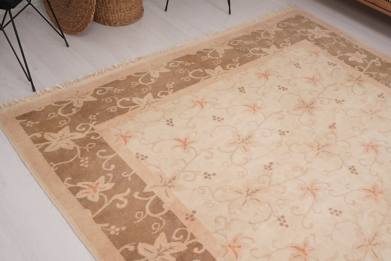 May include: A beige area rug with a brown border. The rug has a floral pattern with a repeating vine design. The border has a similar floral pattern.