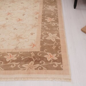 May include: A beige rug with a brown border. The rug has a floral pattern with a repeating design of leaves and vines. The rug has a fringe border.