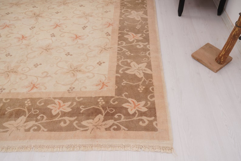 May include: A beige rug with a brown border. The rug has a floral pattern with a repeating design of leaves and vines. The rug has a fringe border.
