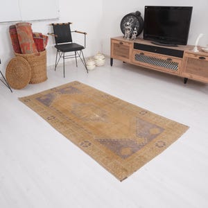 Puede incluir: Una alfombra rectangular de estilo vintage con un diseño central de medallón. La alfombra presenta un campo marrón dorado cálido con una apariencia descolorida y desgastada. El borde y los elementos centrales del diseño son de color púrpura apagado.