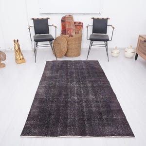Puede incluir: Una alfombra rectangular gris oscuro con una apariencia desgastada y vintage. La alfombra est&aacute; colocada sobre un suelo blanco, con dos sillas negras, un mueble de madera y objetos decorativos en el fondo. Las dimensiones de la alfombra no son visibles.