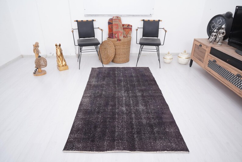 Puede incluir: Una alfombra rectangular gris oscuro con una apariencia desgastada y vintage. La alfombra est&aacute; colocada sobre un suelo blanco, con dos sillas negras, un mueble de madera y objetos decorativos en el fondo. Las dimensiones de la alfombra no son visibles.