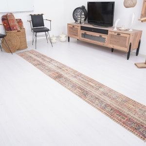May include: A long, rectangular, cream-colored rug with a multi-colored geometric pattern. The rug is laid out on a white floor in a room with furniture.