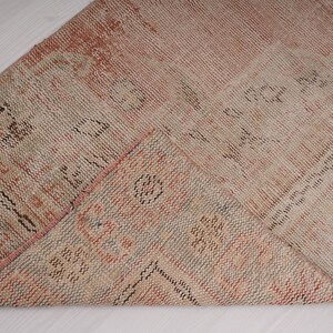 May include: A close-up of a folded, woven rug with a beige and pink color scheme. The rug has a geometric pattern with black accents.